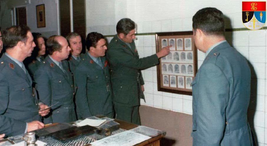 Guardias civiles de uniforme atendiendo a una explicación ante un cuadro de reseñas en una clase de los años 70