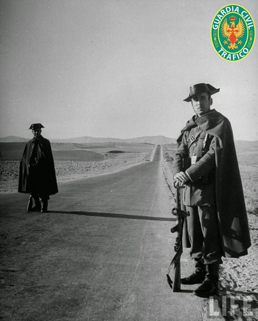 Dos guardias civiles con capa y tricornio vigilando una carretera recta en blanco y negro hacia 1950