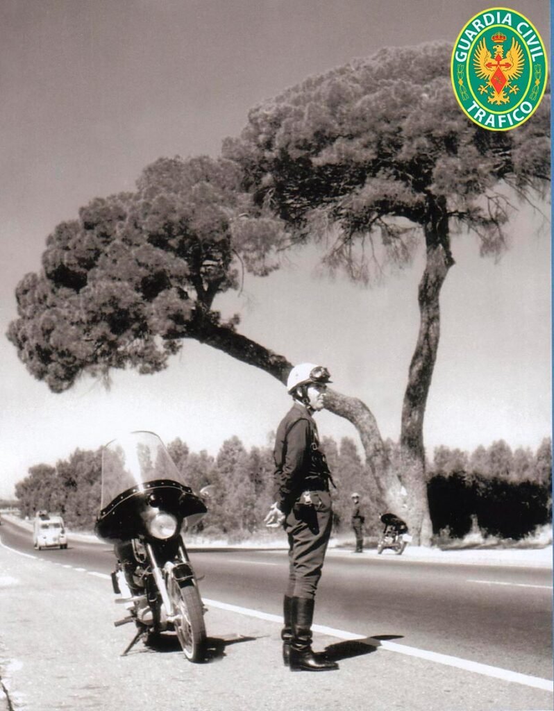 Motorista de la Agrupación de Tráfico junto a su motocicleta bajo un pino, en una carretera de los años 70